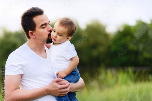Father holding in arms his little son and kissing him outdoor. Green background. Happy family and father care concept. Room for copy text