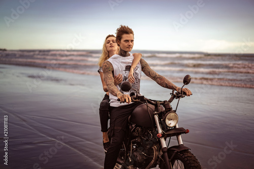 young stylish tattooed couple riding motorcycle on ocean beach
