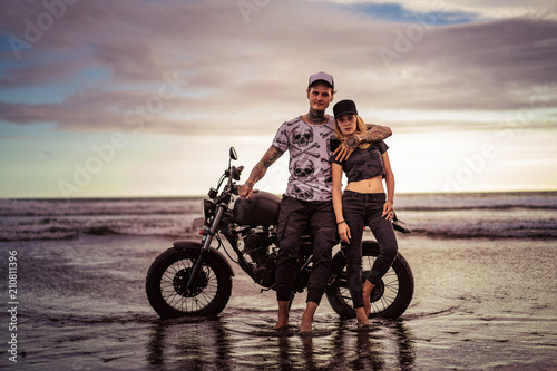 stylish boyfriend hugging girlfriend near motorcycle on ocean beach and they looking at camera