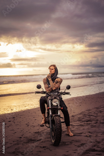 pensive handsome tattooed biker sitting on motorcycle on ocean beach