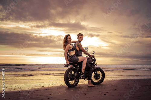 boyfriend and girlfriend sitting on motorcycle at beach during sunrise and looking at camera