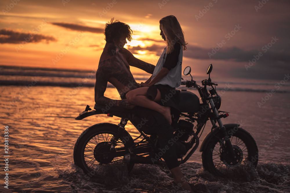 Fototapeta girlfriend sitting on shirtless boyfriend on motorbike at beach during sunset