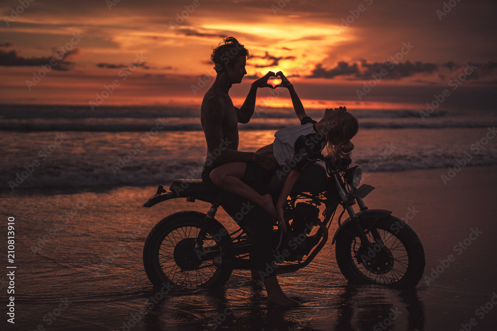 Fototapeta passionate couple making heart with fingers on motorcycle at beach during sunset