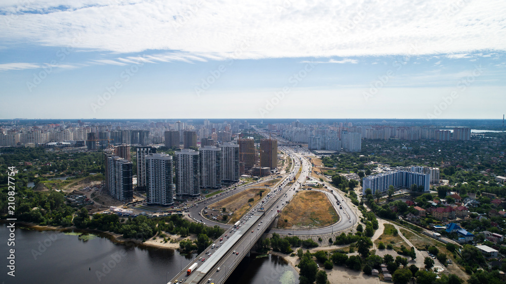 South Bridge. Aerial view of many buildings. The Dnieper River. Summer. The sun. Day. Green trees. High-rise buildings. Road. Cars. Infrastructure. Sky. The clouds. Kyiv.