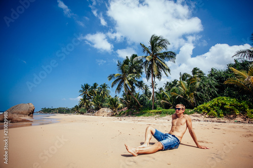 happy holidays in beautiful paradise beach, man sitting on sand beach near ocean and palm trees in shorts and sunglasses and relaxing. Vacation concept.