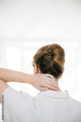 A woman with a fancy updo is touching her neck. She is seen from the back and is wearing a short sleeved beige blouse. photo