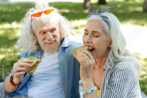 Delicious meal. Delighted positive woman biting a sandwich while enjoying the meal together with her husband