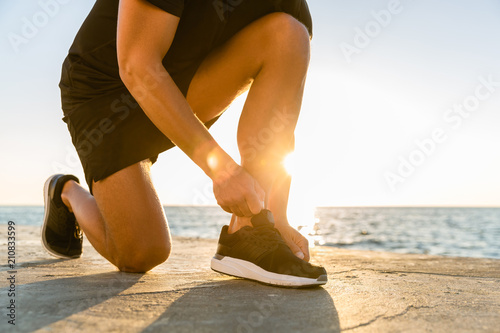 cropped shot of sportsman lacing up sneakers on seashore in front of sunrise
