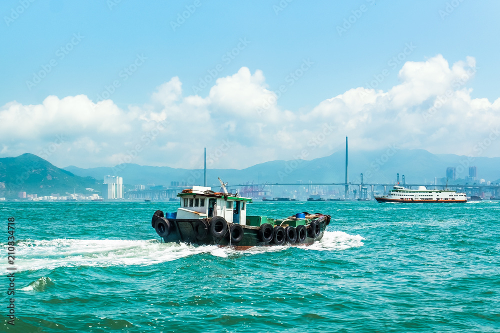 Obraz premium The tugboat passing across Victoria Harbour, Hong Kong. Beautiful aquamarine sea water, blue sky with white clouds. Bridge, cargo container port and terminal on background.