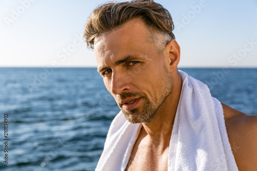 shirtless handsome adult man with towel on shoulders looking at camera on seashore