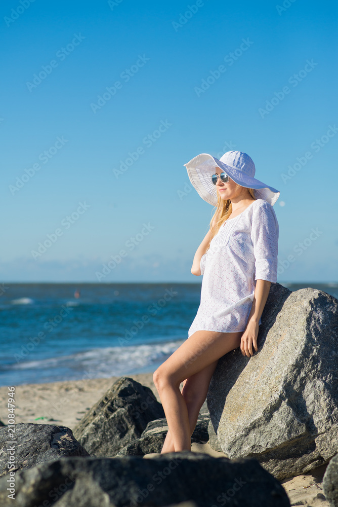 Attractive woman in white dress, big hat and glasses relaxing on the beach on sunny day