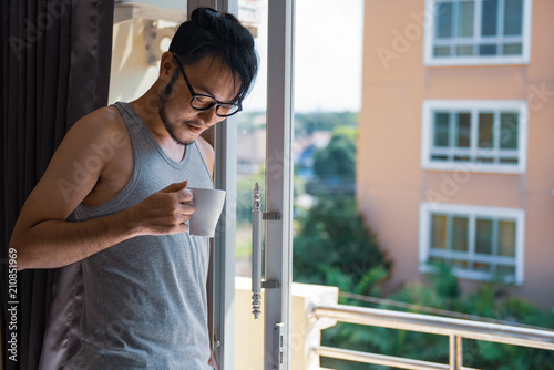 Young asian man having a coffee at the balcony glasses window in the wonderful day morning.