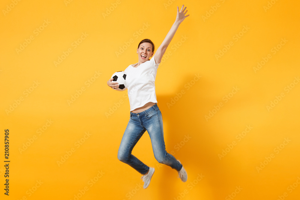 Young fun expressive European woman football fan jumping in air, cheer up support team, holding soccer ball isolated on yellow background. Sport, play football, cheer, fans people lifestyle concept.