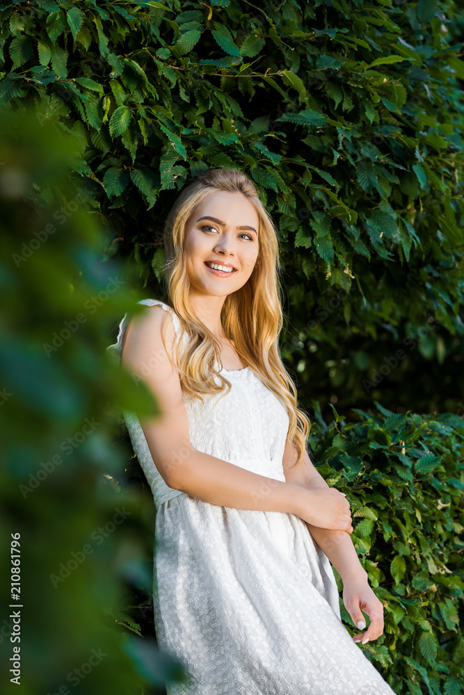 selective focus of beautiful smiling blonde girl in white dress posing near green leaves selective focus of beautiful smiling blonde girl in white dress posing near green leaves