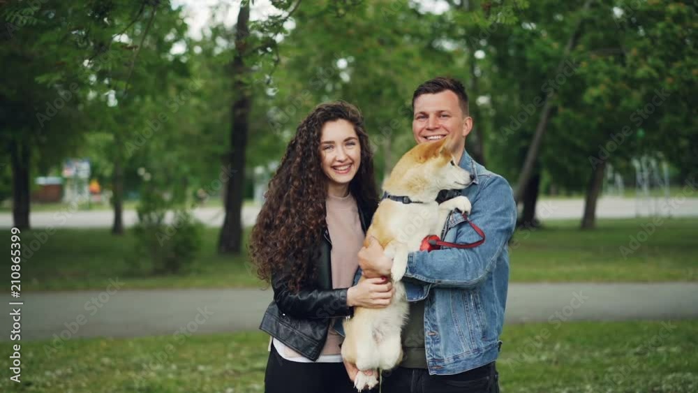 Slow motion portrait of man and woman proud dog owners holding adorable puppy, looking at camera and smiling standing in park on summer day. Slow motion portrait of man and woman proud dog owners holding adorable puppy, looking at camera and smiling standing in park on summer day.