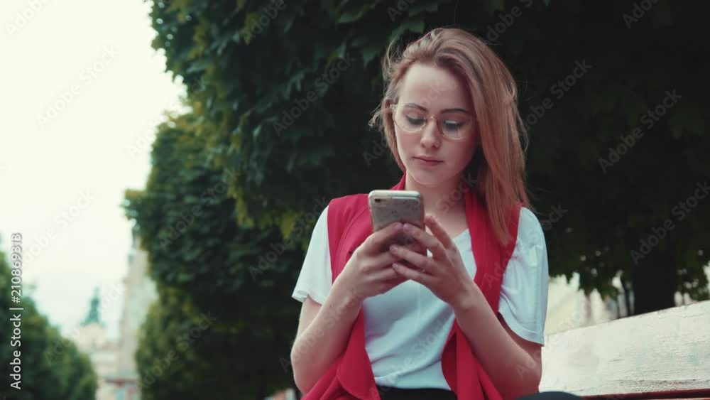 Serious pretty young woman in trendy casual outfit sits on a bench in the city park, actively texts messages via her phone. Being online, modern technologies, popularity. Addiction, gadgets, devices. Serious pretty young woman in trendy casual outfit sits on a bench in the city park, actively texts messages via her phone. Being online, modern technologies, popularity. Addiction, gadgets, devices.