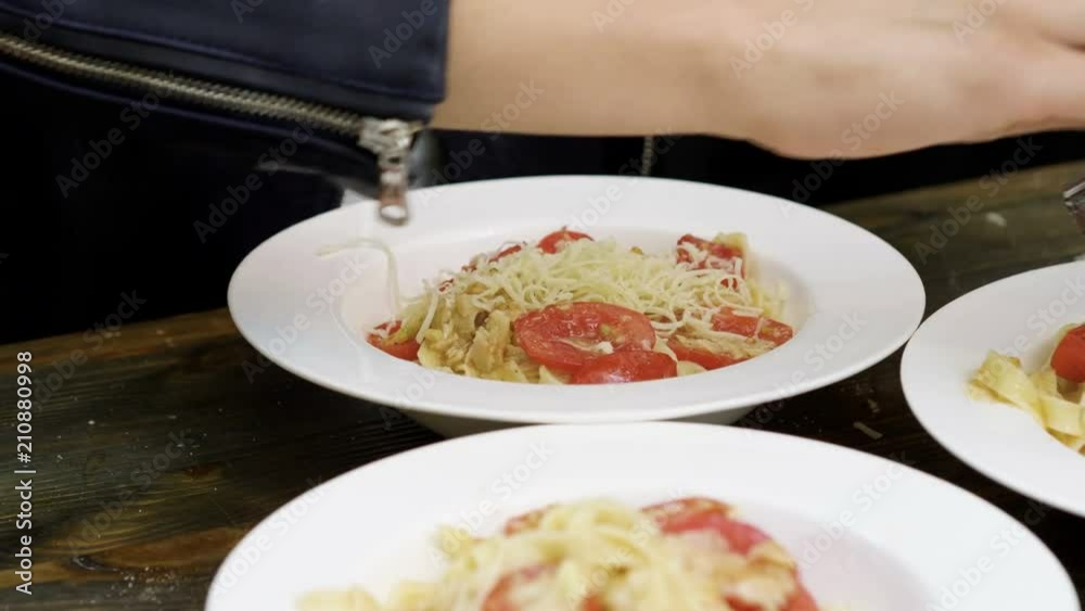 Women's hands rubbing parmesan cheese on a cooked pasta with tomatoes, almonds and pesto sauce on a white plate. 4K