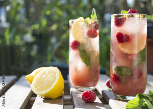 Raspberry ice tea in tall glass with fresh raspberries and lemon. Served on wooden table in a flower garden