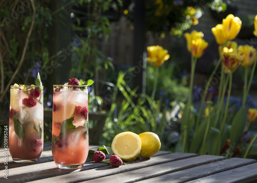 Raspberry ice tea in tall glass with fresh raspberries and lemon. Served on wooden table in a flower garden