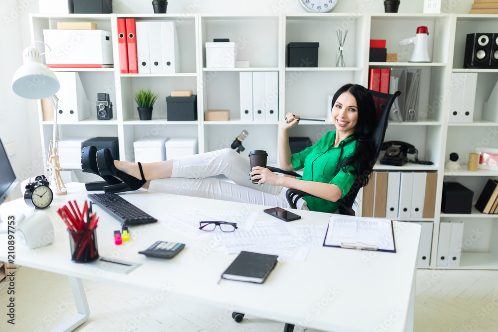 Obraz premium A young girl sits in the office, threw her legs on the table and holds a glass of coffee.