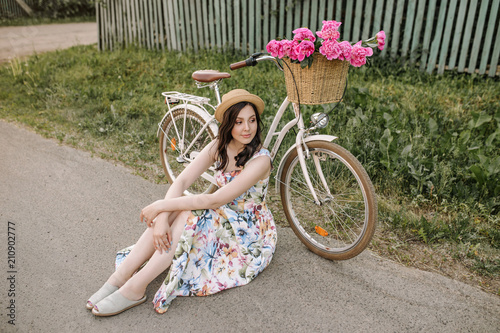 Smiling cute happy girl in dress and hat is seatng near her retro white bicycle with flowers in the buncket. Green sunny park is around photo