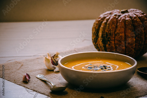 Pumpkin soup with pumpkin seeds. In white bowl, on hessian fabric. With silver spoon, garlic and whole pumpkin.. 