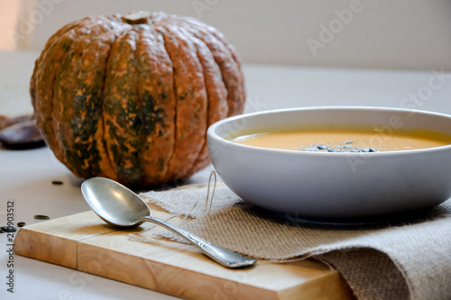 Pumpkin soup with pumpkin seeds. In white bowl, on hessian fabric and wooden board. With silver spoon, whole pumpkin.