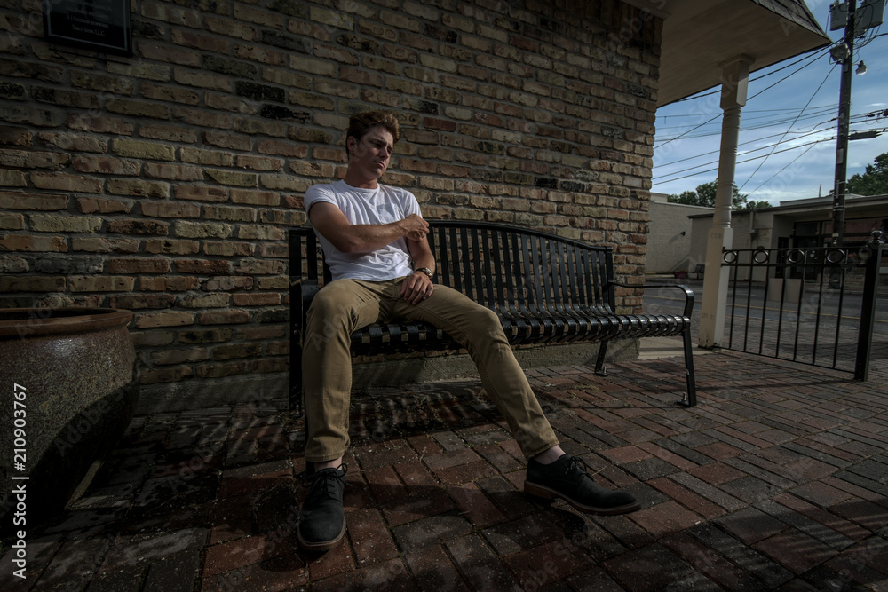 Young man sits on a bench in a retro area of town wearing a white shirt and khaki pants on a nice sunny day.