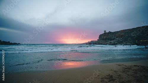 Pink and purple sunset on the beach. Silhouette of the surfer man with the surfboard waiting on the wave in the evening in the sea.