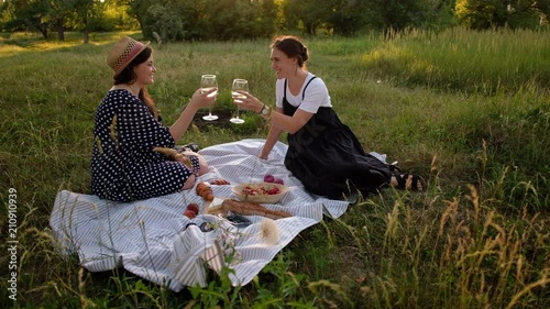 Two young women enjoying a picnic on the grass at warm summer evening during sunset. They are tosting and drinking white wine photo