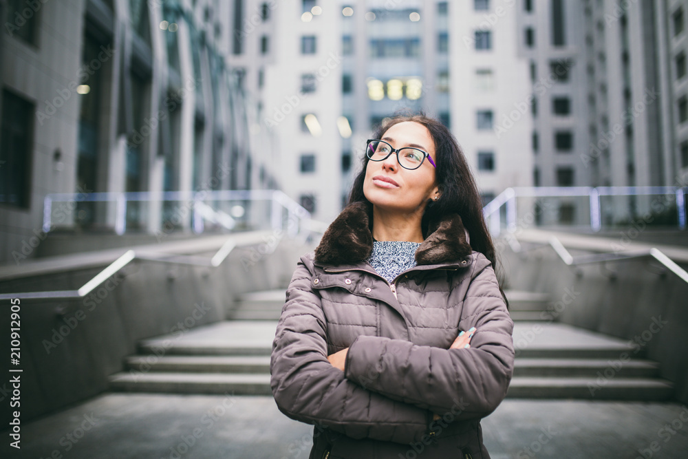 Beautiful young woman of European ethnicity with long brunette hair looking to side and up, wearing glasses and coat stands on background of the business center in the fall in cloudy cloudy weather