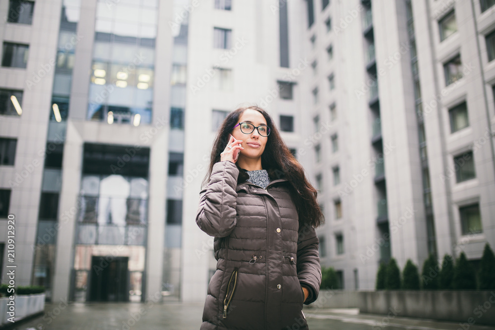 Theme is the business situation. Beautiful young woman of European ethnicity with long brunette hair wearing glasses and coat stands on background of business center and uses phone in hand near ear