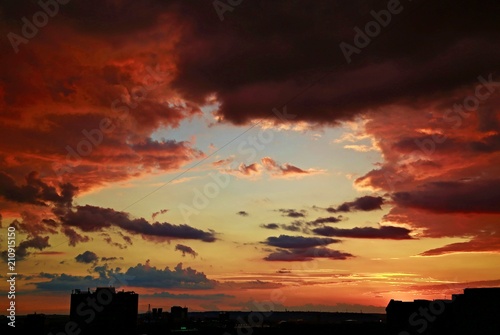 Dark colorful dramatic sky with purple and pink clouds, red and golden sunset behind houses, black horizon with city landscape silhouette, blue window into heaven, contrast colors