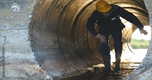 Engineer, worker checks gutters, in blue robe, in boots, in a construction yellow helmet, tunnels, technical supervision.