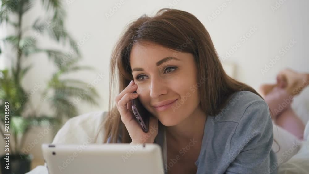 Face on beautiful woman lying on the bed and having phone talk with plant on the background. Indoors. Portrait. Face on beautiful woman lying on the bed and having phone talk with plant on the background. Indoors. Portrait.