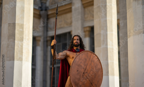 a Spartan warrior with a spear and shield. buildings with columns on the background.
