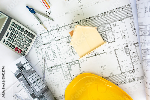 Wooden house model on engineering blueprint with architecture drawing plan, caculator, circus, pencil and yellow helmet.