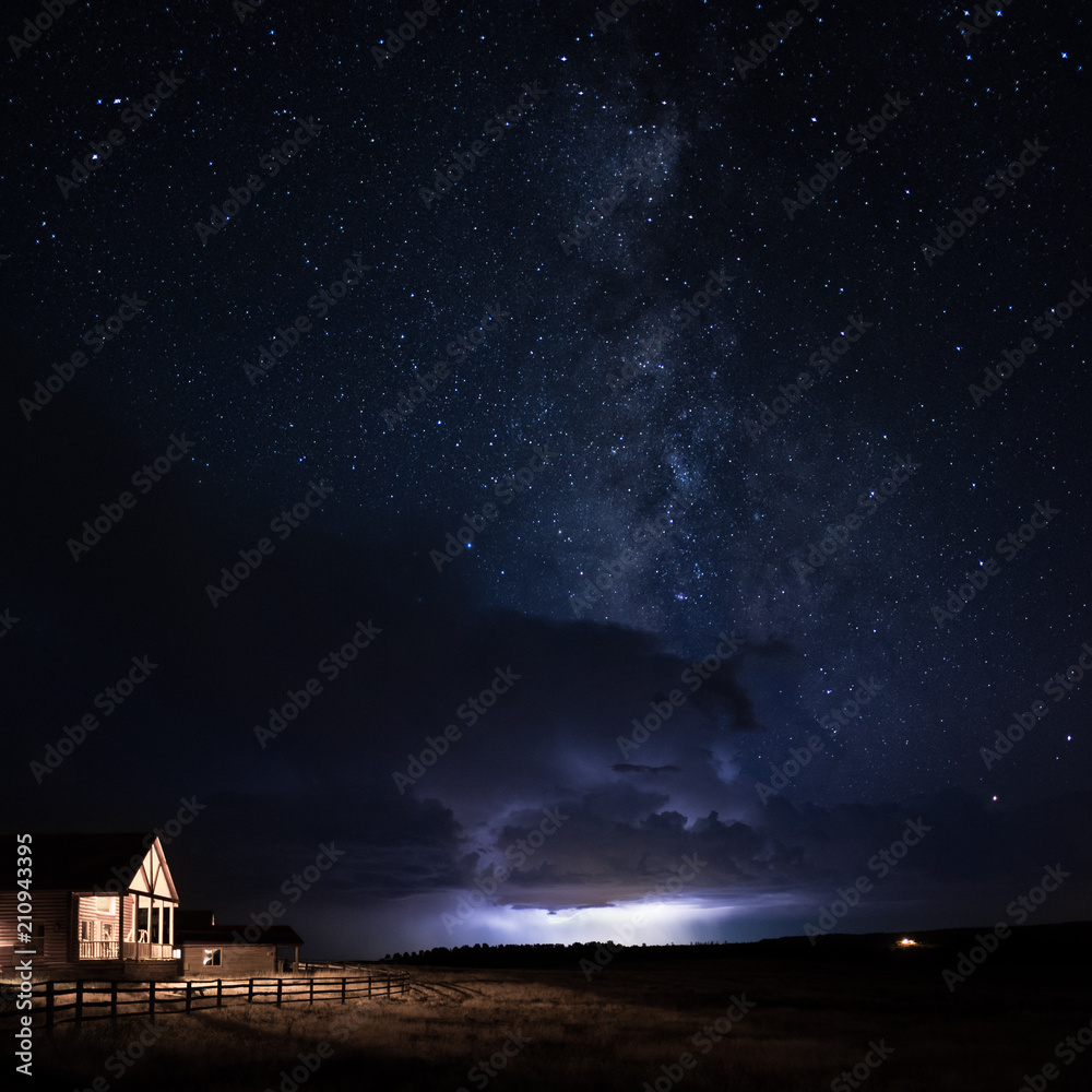 A wood cabin with the porch light on with a wooden fence and grass field with a lightning storm and dramatic clouds behind it with the Milky Way galaxy rising above it in a starry night sky A wood cabin with the porch light on with a wooden fence and grass field with a lightning storm and dramatic clouds behind it with the Milky Way galaxy rising above it in a starry night sky