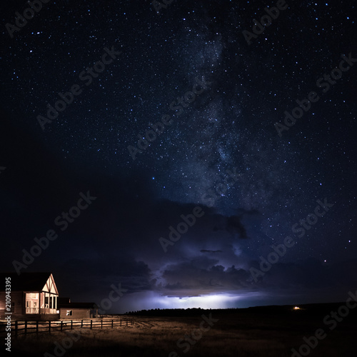 Wallpaper Mural A wood cabin with the porch light on with a wooden fence and grass field with a lightning storm and dramatic clouds behind it with the Milky Way galaxy rising above it in a starry night sky Torontodigital.ca