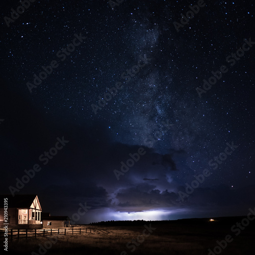 A wood cabin with the porch light on with a wooden fence and grass field with a lightning storm and dramatic clouds behind it with the Milky Way galaxy rising above it in a starry night sky