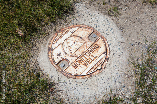 Old Decayed Monmitoring Well Lid Set in Concrete, Surrounded by Soil and Grass