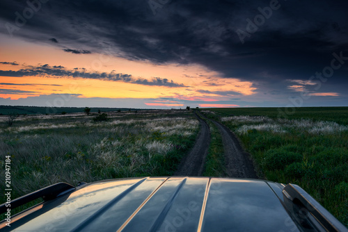 the car rides on a dirt road in the field, beautiful sunset with wild grass, sunlight and dark clouds