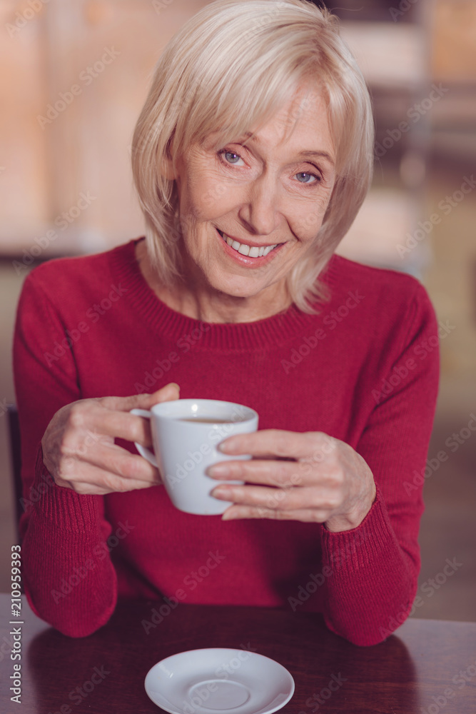 Sincere smile. Delighted blonde holding cup in both hands while drinking coffee Sincere smile. Delighted blonde holding cup in both hands while drinking coffee