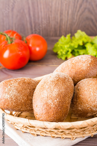 Fresh rye buns in a basket, salad and tomato on a wooden table