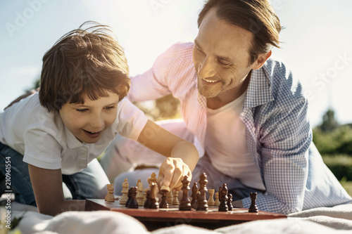 Favourite game. Joyful laughing kneeling boy playing chess with his dad enjoying the nature and his dad smiling