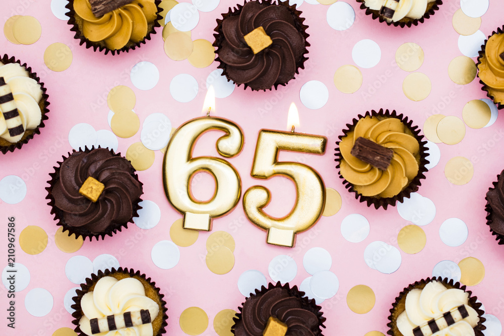 Number 65 gold candle with cupcakes against a pastel pink background Number 65 gold candle with cupcakes against a pastel pink background