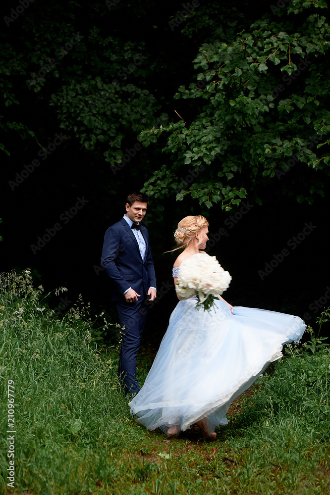The bride is spinning, in a blue beautiful dress. The groom stands at a distance and admires. Park or forest. Wedding walk and photo shoot The bride is spinning, in a blue beautiful dress. The groom stands at a distance and admires. Park or forest. Wedding walk and photo shoot