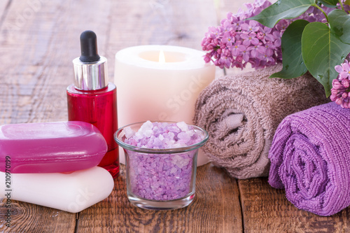 Red bottle with aromatic oil  burning candle  soap  bowl with sea salt  lilac flowers and towels
