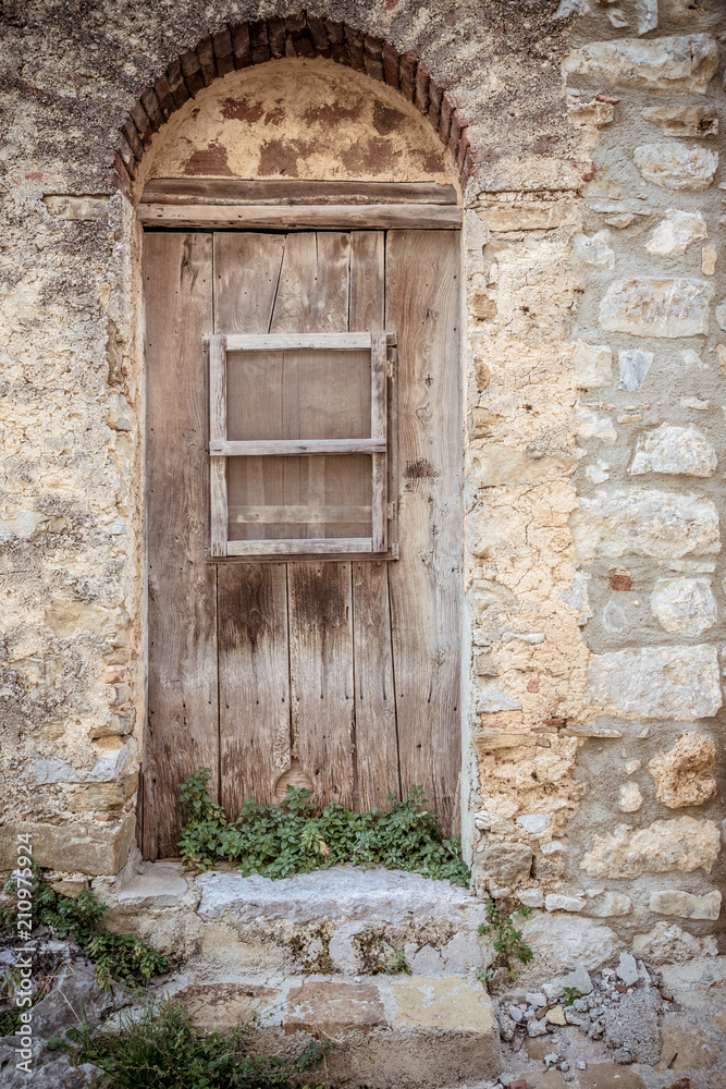 Old brown wooden door and dirty stone wall. Plants growing and two stone steps. Entrance to the house Old brown wooden door and dirty stone wall. Plants growing and two stone steps. Entrance to the house