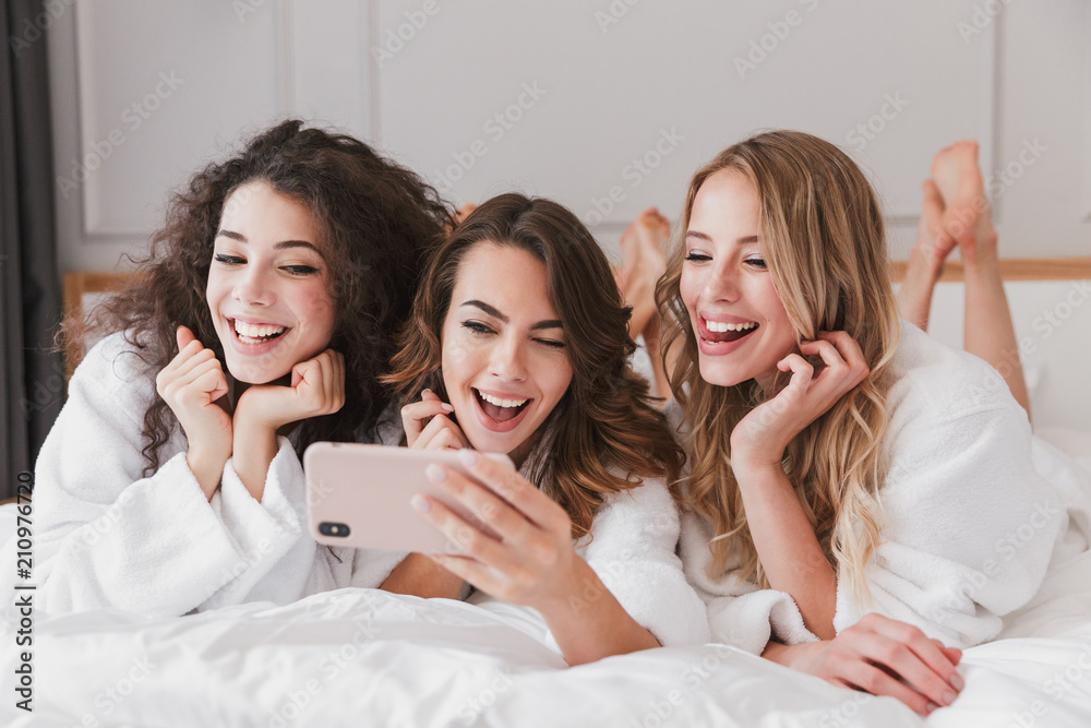 Happy photo of lovely smiling women 20s wearing white bathrobe lying in luxury bedroom in hotel room, and taking selfie on cell phone during hen party Happy photo of lovely smiling women 20s wearing white bathrobe lying in luxury bedroom in hotel room, and taking selfie on cell phone during hen party